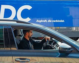 Ein Mann fährt mit einem Auto an einer blauen Lok mit RDC-Schriftzug vorbei.