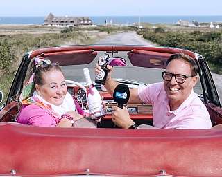 Frank Bremser und Conny Reckert sitzen in mit Getränken in einem Cabrio vor einer Sylter Dünenlandschaft.